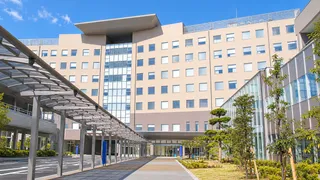 青空の下に建つ深谷赤十字病院と、正面入口へ続く屋根付きの通路のイメージ
